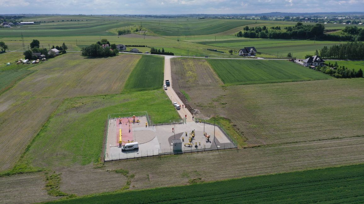 Panorama z lotu ptaka przedstawiająca prostokątny, ogrodzony obiekt gazowy (zespół zaporowo-upustowy) w rolniczym krajobrazie. Do stacji prowadzi utwardzona droga dojazdowa, na której zaparkowane są białe pojazdy techniczne. Wokół instalacji rozciągają się wielobarwne, podłużne pasy pól uprawnych tworzące regularną mozaikę. Na dalszym planie widać pojedyncze gospodarstwa domowe.