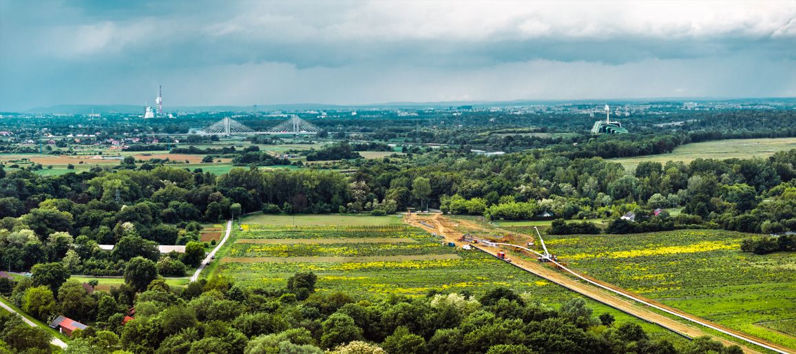 Panoramiczne zdjęcie lotnicze ukazujące pas budowy gazociągu przecinający rozległe zielone oraz żółte, kwitnące pola. Wokół widoczne są liczne kępy drzew i zielone zagajniki. Na dalekim planie, pod sinym, burzowym niebem, rozciąga się miejska panorama z charakterystycznym białym mostem wantowym oraz wysokim kominem elektrociepłowni.