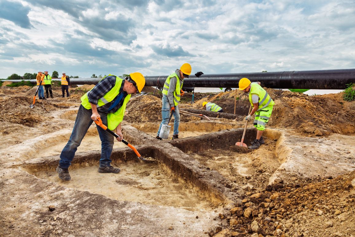 Zdjęcie z poziomu gruntu przedstawiające grupę archeologów pracujących w głębokich, regularnych wcięciach w ziemi. Mężczyźni w żółtych kaskach i kamizelkach odblaskowych ostrożnie oczyszczają łopatami odsłonięte profile i jamy. W tle, na podwyższeniu terenu, przebiega zamontowana, potężna czarna rura gazociągu. Nad całością rozciąga się zachmurzone niebo.