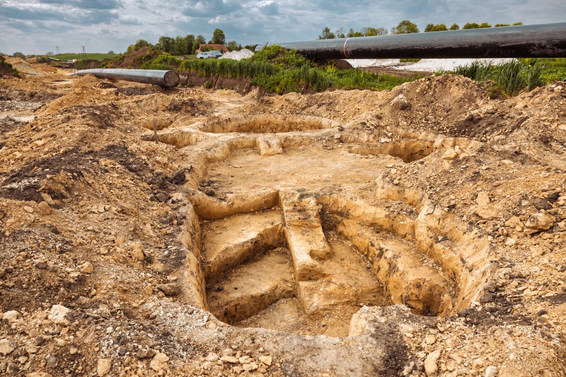 Zdjęcie przedstawia zbliżenie na stanowisko archeologiczne po usunięciu wierzchniej warstwy ziemi. W jasnej, gliniastej glebie widoczne są misternie doczyszczone, schodkowe struktury i okrągłe wcięcia będące dawnymi obiektami ziemnymi. W tle, na krawędzi wykopu, ciągnie się czarna nitka gazociągu, za którą widać pas zieleni i zaparkowane samochody.