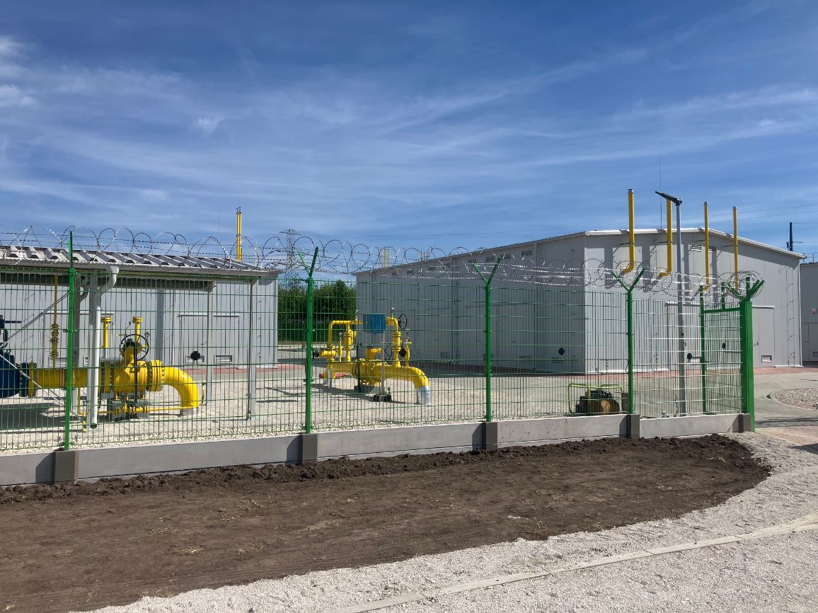 Zdjęcie z poziomu gruntu przedstawiające infrastrukturę technologiczną. Za zielonym płotem panelowym zwieńczonym drutem ostrzowym (żyletkowym) widoczne są potężne, jaskrawożółte instalacje rurowe, zasuwy oraz zawory gazowe posadowione na żwirowym podłożu. W tle stoją dwa nowoczesne, industrialne budynki modułowe o jasnoszarych ścianach. Całość sfotografowana na tle bezchmurnego, niebieskiego nieba.