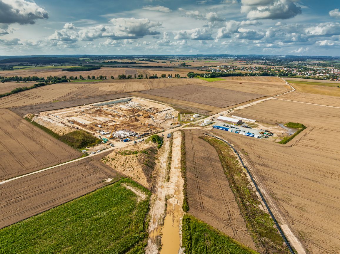 Widok z lotu ptaka na rozległy plac budowy lądowej infrastruktury gazowej, otoczony mozaiką brązowych i zielonych pól uprawnych. Na terenie inwestycji widoczne są wykopy, powstające fundamenty, drogi technologiczne oraz zaplecze kontenerowe pod niebem z białymi chmurami.