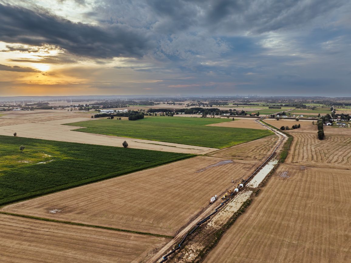 Panoramiczny widok z lotu ptaka na pas budowy rurociągu lądowego, który przecina rozległe, wielobarwne pola uprawne i zmierza w kierunku horyzontu. Na trasie widoczne są miniaturowe sylwetki maszyn budowlanych. Całość sfotografowana w świetle zachodzącego słońca z dramatycznymi chmurami.