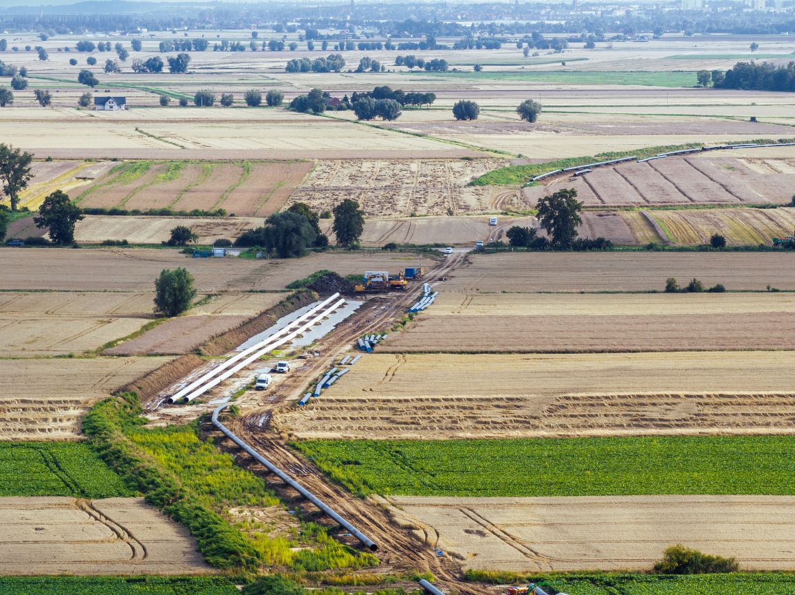 Szeroka panorama z lotu ptaka na rozległy krajobraz rolniczy z geometryczną mozaiką zielonych i złotych pól. Przez środek kadru przebiega linia budowy gazociągu z widocznymi maszynami i rurami. Na dalekim horyzoncie rysuje się zamglona sylwetka miasta.
