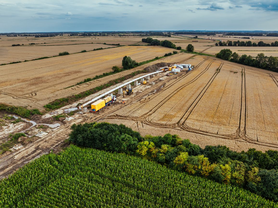 Zdjęcie z drona przedstawiające pas instalacji rurociągu biegnący między zżętym polem zboża a zieloną uprawą kukurydzy. Długa, biała rura spoczywa na podwyższeniach. Na trasie prac widoczna jest ciężarówka z żółtym kontenerem, koparka oraz pracownicy w kamizelkach odblaskowych.