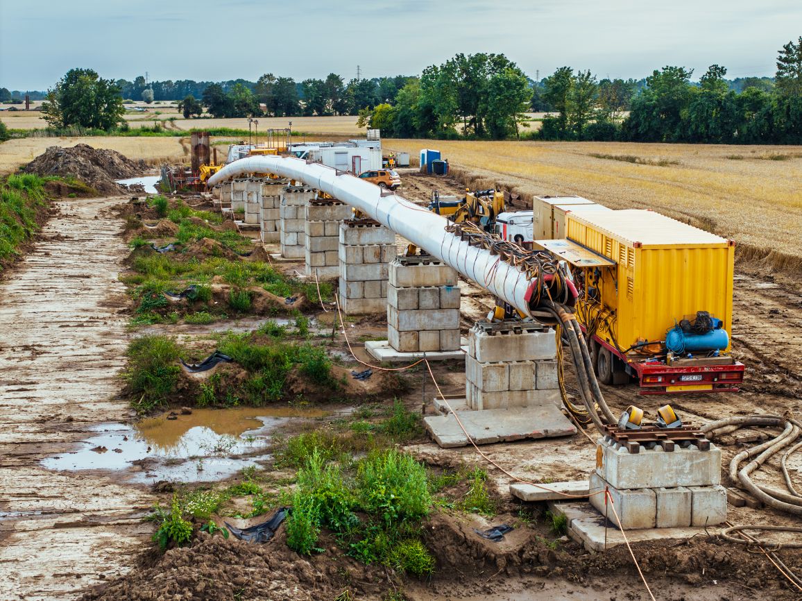 Zdjęcie z poziomu ziemi przedstawiające odcinek grubego, białego rurociągu zainstalowanego na rzędzie masywnych, tymczasowych podpór wykonanych z betonowych bloków. Po prawej stronie stoi duża, żółta maszyna przemysłowa (kontener technologiczny) na czerwonym podwoziu, z której do wnętrza rury poprowadzono wiązki grubych, czarnych przewodów i rur strumieniowych. Teren wokół jest błotnisty z kałużami, a w tle widać pole dojrzewającego zboża oraz ścianę zielonych drzew.