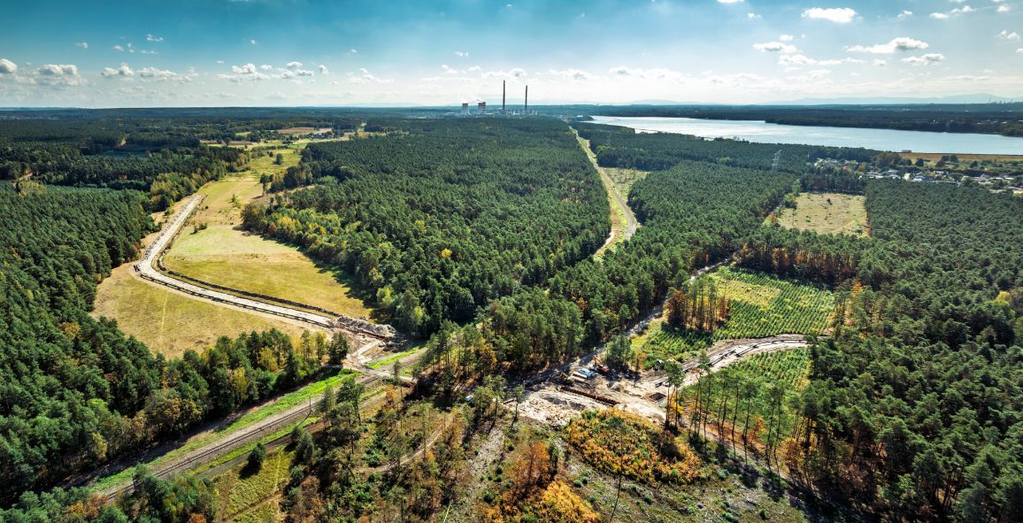Widok lotniczy na teren budowy gazociągu przebiegający przez rozległy kompleks leśny. Wśród drzew widoczny jest szeroki, wykarczowany pas terenu z drogami technicznymi i pracami ziemnymi związanymi z układaniem rurociągu. W tle znajduje się duży zbiornik wodny oraz obiekty przemysłowe z wysokimi kominami. Zdjęcie wykonano w słoneczny dzień przy dobrej widoczności.