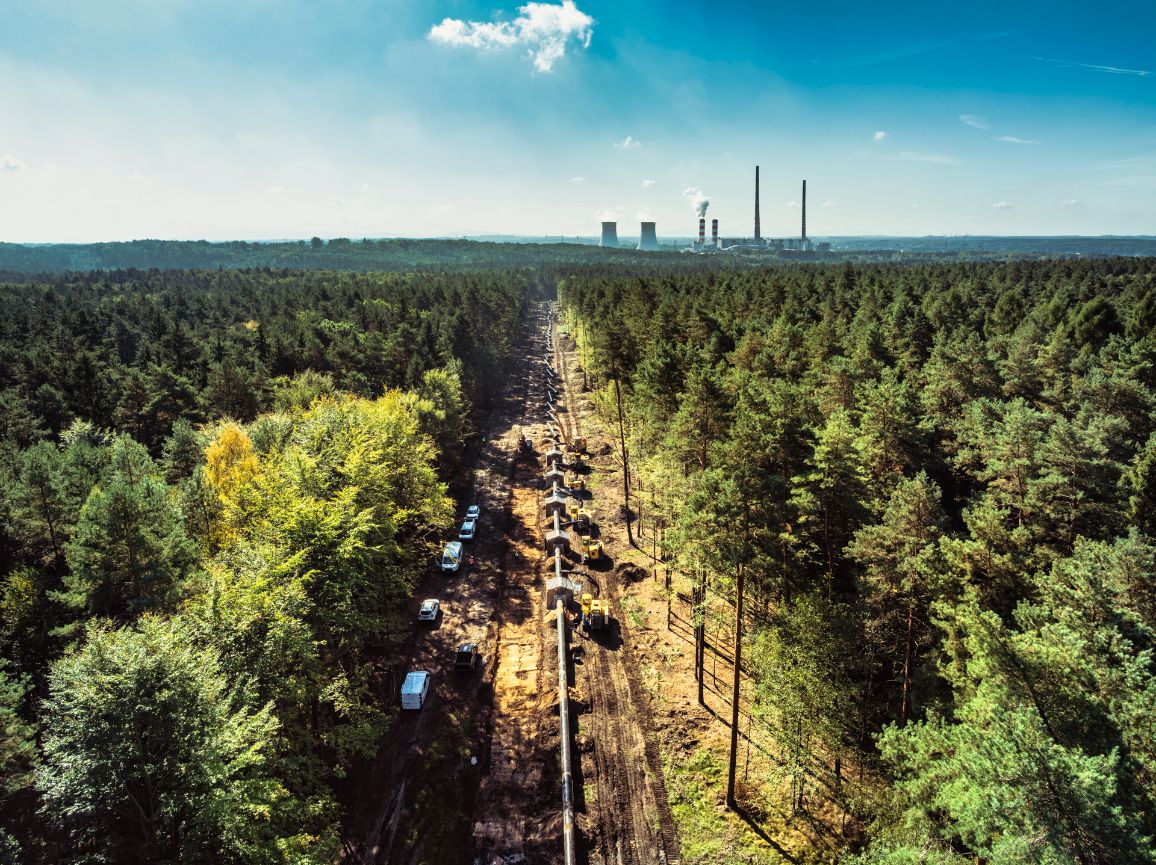 Ujęcie z góry na długą, prostą przecinkę w lesie, wzdłuż której układana jest linia gazociągu. Na trasie stoją zaparkowane maszyny budowlane oraz białe samochody serwisowe, a na odległym horyzoncie majaczą chłodnie kominowe i kominy elektrowni.