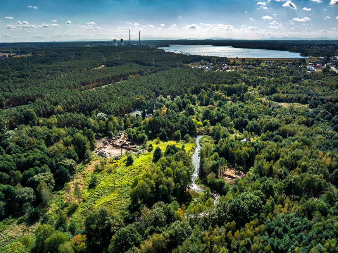 Widok z lotu ptaka na zielony krajobraz z zakolem rzeki i placem budowy gazociągu w środku lasu. W tle widoczny jest duży, oświetlony słońcem zbiornik wodny oraz zabudowania przemysłowe elektrowni.