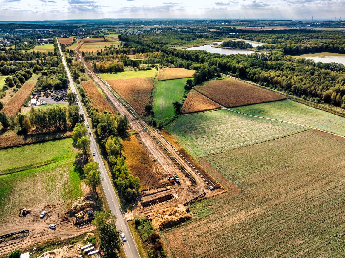 Widok z lotu ptaka na pas budowy gazociągu biegnący równolegle do lokalnej drogi asfaltowej. Na placu robót pośród pól uprawnych widać głęboki wykop, ułożone w rzędzie rury oraz maszyny budowlane. W tle widoczne są stawy i kompleks leśny.