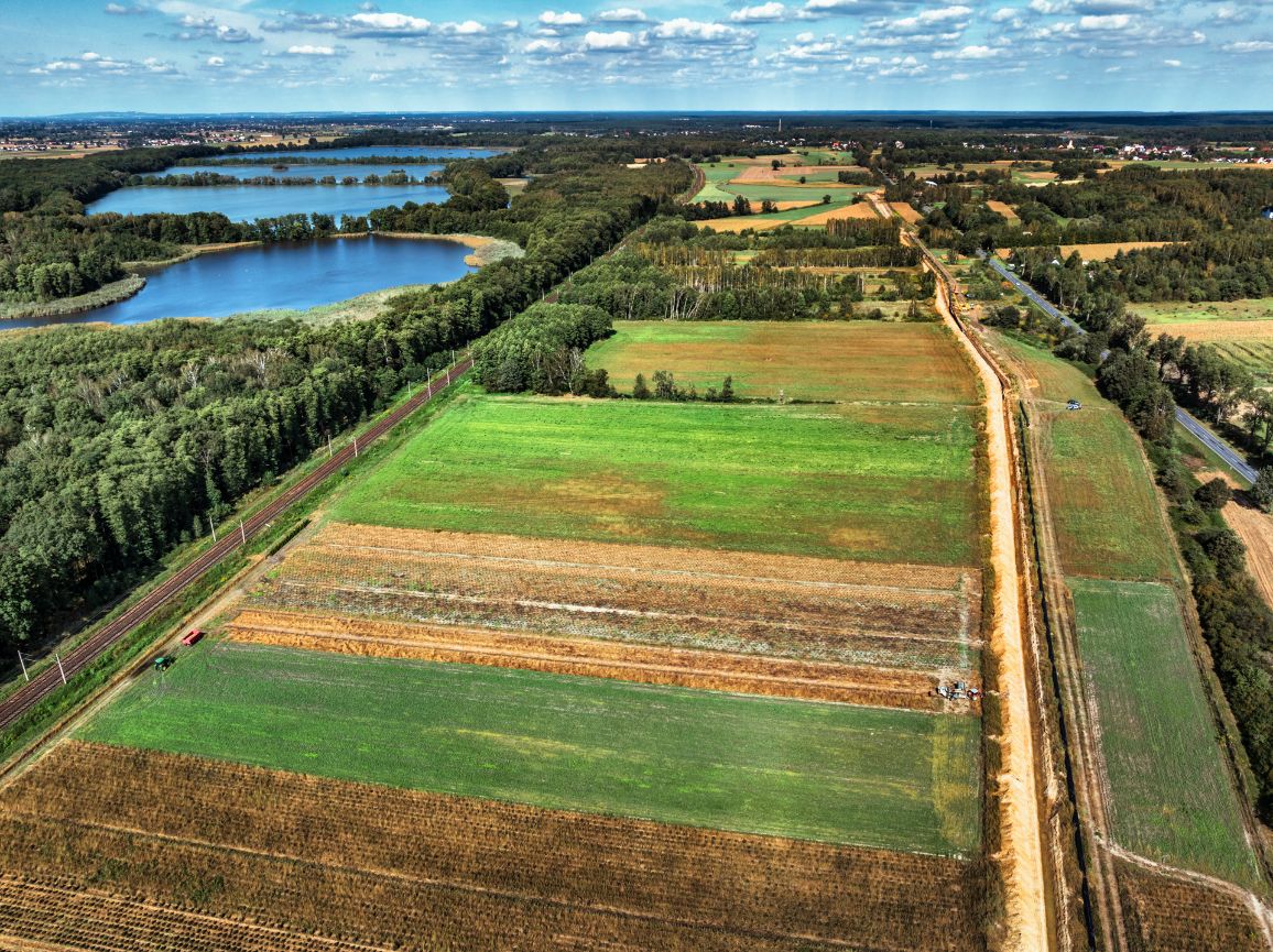 Widok z lotu ptaka na prosty, piaszczysty pas inwestycji gazowej graniczący z polami uprawnymi. Po lewej stronie przebiega linia kolejowa oddzielająca plac budowy od gęstego lasu, za którym widać dwa jeziora.