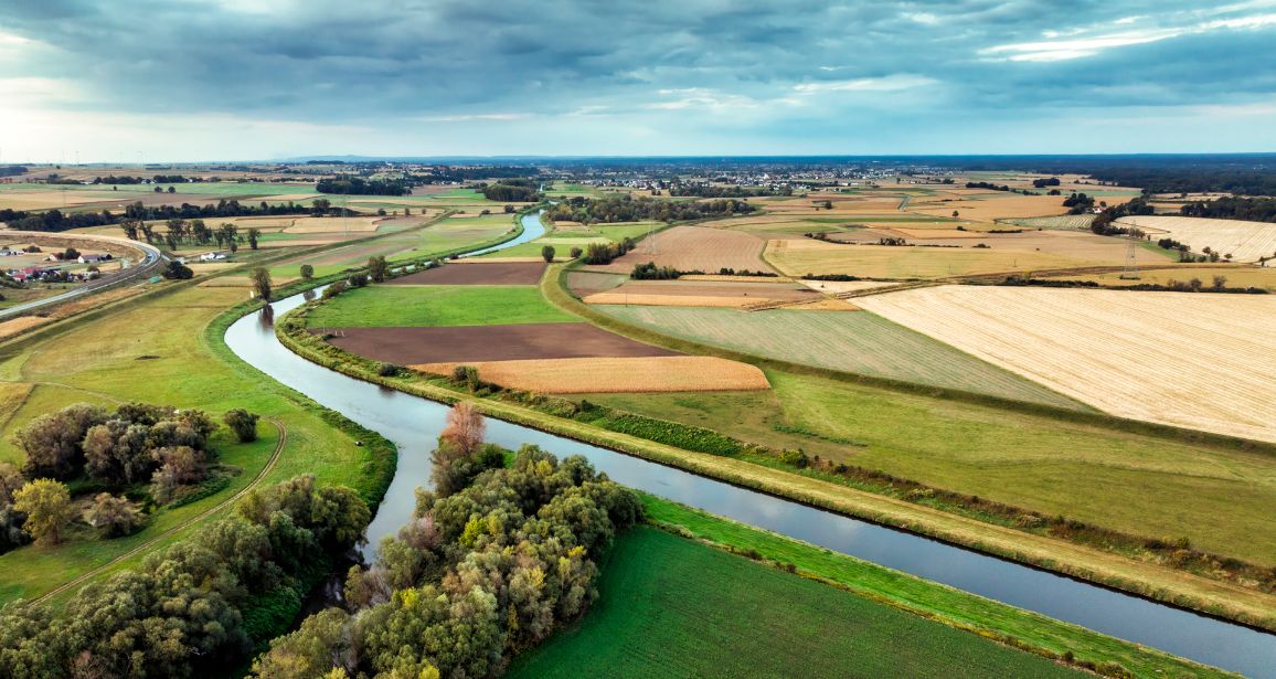 Panoramiczny widok z lotu ptaka na malowniczą dolinę z zakolami rzeki. Rzeka meandruje pomiędzy różnobarwnymi, geometrycznymi polami uprawnymi i kępami drzew pod częściowo zachmurzonym niebem.