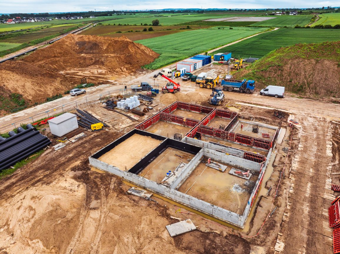 Widok z lotu ptaka na plac budowy lądowej części inwestycji FSRU, gdzie wznoszone są fundamenty nowego obiektu. Widoczna jest betonowa konstrukcja z czerwonymi szalunkami ściennymi, otoczona przez maszyny budowlane, samochody ciężarowe, kontenery pracownicze i pryzmy wykopanej ziemi