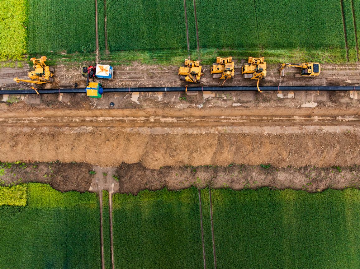 Pionowe ujęcie bezpośrednio z góry na zaawansowane prace ziemne lądowej części inwestycji FSRU. Wzdłuż wykopanego w ziemi głębokiego, prostego rowu ustawiony jest równolegle rząd żółtych maszyn budowlanych, które podtrzymują długą nitkę stalowego rurociągu biegnącego przez zielone pola.