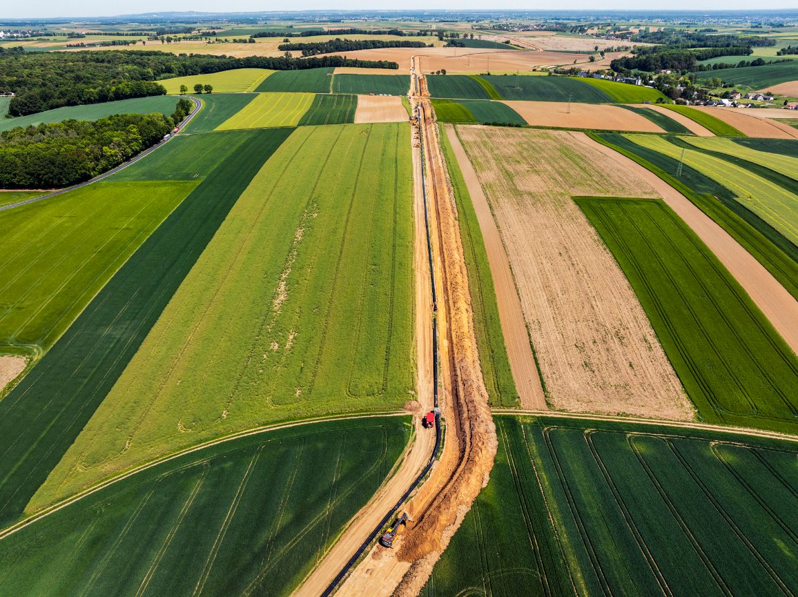 Wysokie ujęcie z lotu ptaka na prosty pas budowy gazociągu, biegnący przez środek kolorowej mozaiki wielkich pól uprawnych. Wzdłuż wykopu z widoczną brunatną ziemią ciągnie się czarna nitka rurociągu, przy której stoi pojedynczy czerwony pojazd.