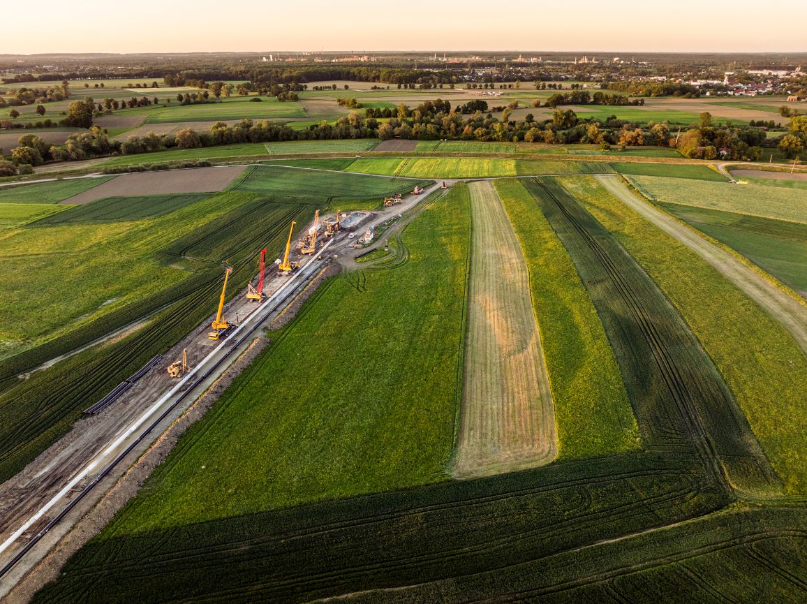 Szeroka panorama lotnicza placu montażowego zorganizowanego na zielonym polu w blasku zachodzącego słońca. W równej linii wzdłuż białego odcinka rury stoją zaparkowane ciężkie dźwigi samojezdne z podniesionymi wysięgnikami.