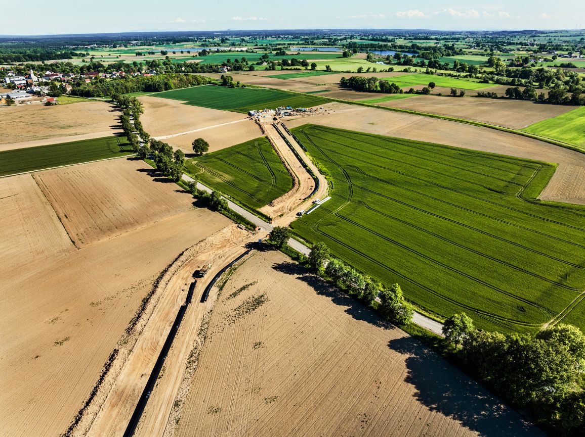 Widok z góry na pas budowy gazociągu, który przecina wąską, asfaltową drogę obsadzoną szpalerem zielonych drzew. Piaszczysta trasa inwestycji łączy brązowe, zaorane pole na pierwszym planie z dużym, zielonym polem po drugiej stronie drogi.