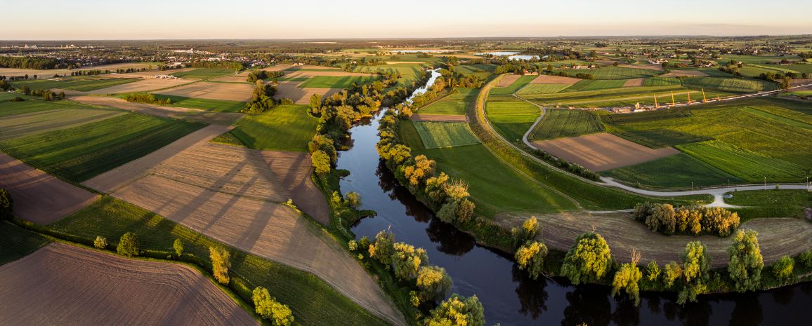 Szeroka panorama doliny rzeki wijącej się zakolami wśród malowniczych pól uprawnych w ciepłym świetle zachodzącego słońca. W oddali, po prawej stronie, widoczny jest podłużny plac budowy gazociągu z zapleczem technicznym.