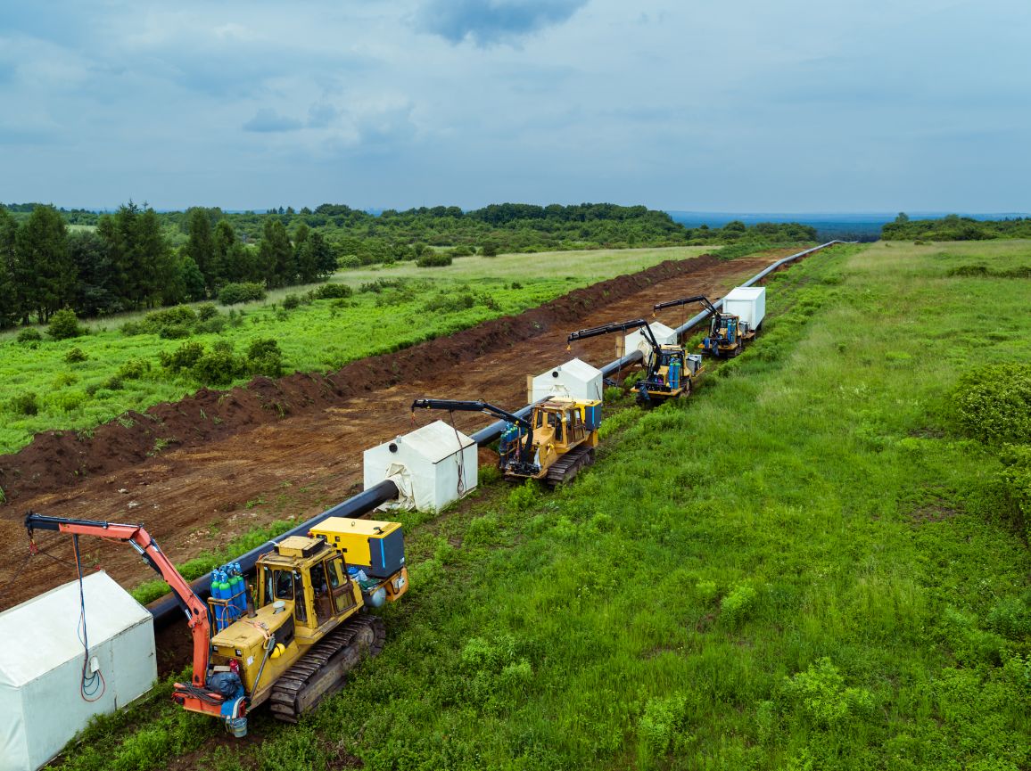 Widok z lotu ptaka na pas montażowy gazociągu przecinający rozległe zielone łąki. Wzdłuż ułożonej na powierzchni ziemi linii czarnych rur stoi kolumna żółtych maszyn budowlanych (układarek bocznych). Nad miejscami łączenia rur rozstawione są białe namioty spawalnicze, chroniące stanowiska pracy przed warunkami atmosferycznymi.