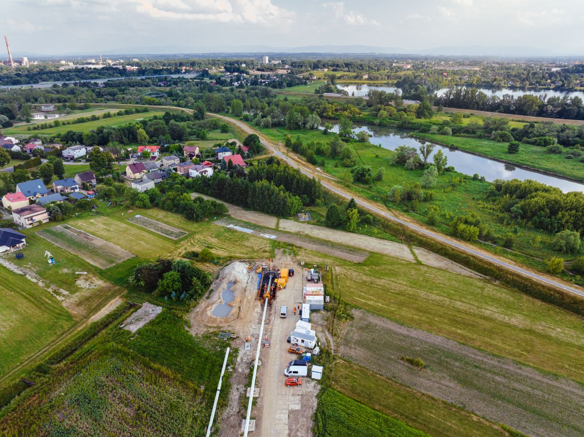 Zdjęcie lotnicze placu budowy zlokalizowanego w pobliżu rzeki. W dolnej części kadru widoczna jest długa, przygotowana do wciągnięcia nitka białego gazociągu, która prowadzi prosto do zabezpieczonej komory startowej przewiertu. Wokół komory zaparkowane są pojazdy techniczne i kontenery. W tle meandruje Wisła otoczona zielenią i zabudowaniami.