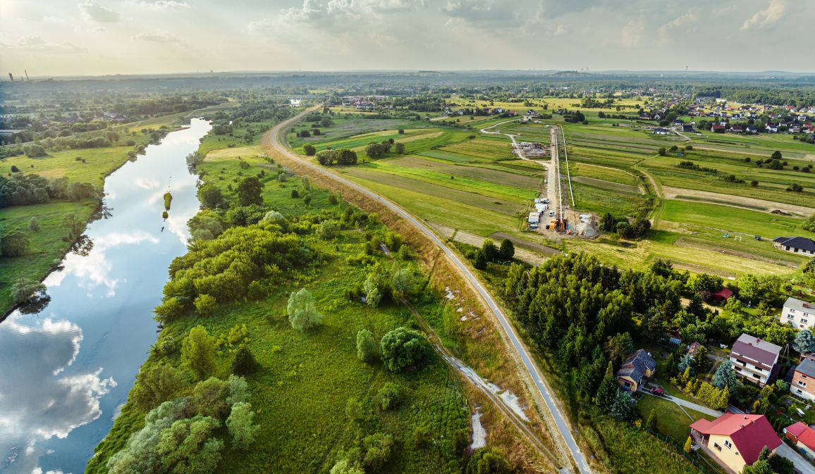 Panoramiczne ujęcie z góry przedstawiające szerokie zakole rzeki Wisły, w której tafli odbijają się chmury. Po prawej stronie rzeki, pośród mozaiki zielonych pól uprawnych, ciągnie się prosta, piaszczysta linia placu budowy gazociągu wraz z widocznym w oddali zapleczem technologicznym przewiertu Direct Pipe.