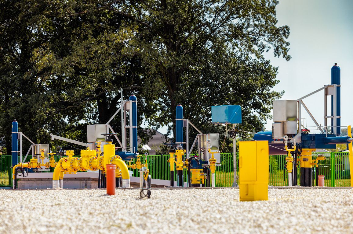 Szeroki kadr na naziemną stację gazową z różnorodną armaturą: żółtymi rurami, niebieskimi cylindrami sterującymi i szarymi skrzynkami elektrycznymi. Plac stacji pokryty jest białym kamieniem, a tło stanowią wysokie, zielone drzewa.
