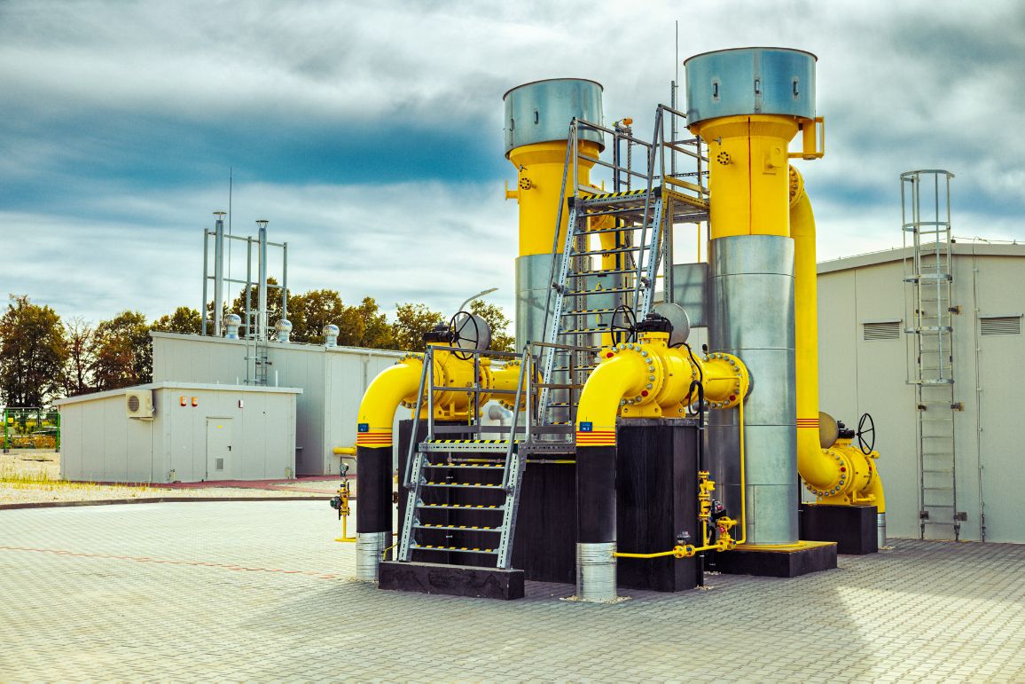 Dwa potężne, pionowe filtry-separatory gazu w kolorze żółtym ze srebrnymi, metalowymi osłonami w górnej części. Do urządzeń prowadzi ocynkowana konstrukcja schodów i pomostów technicznych. W tle widoczny kontenerowy budynek pod zachmurzonym niebem.