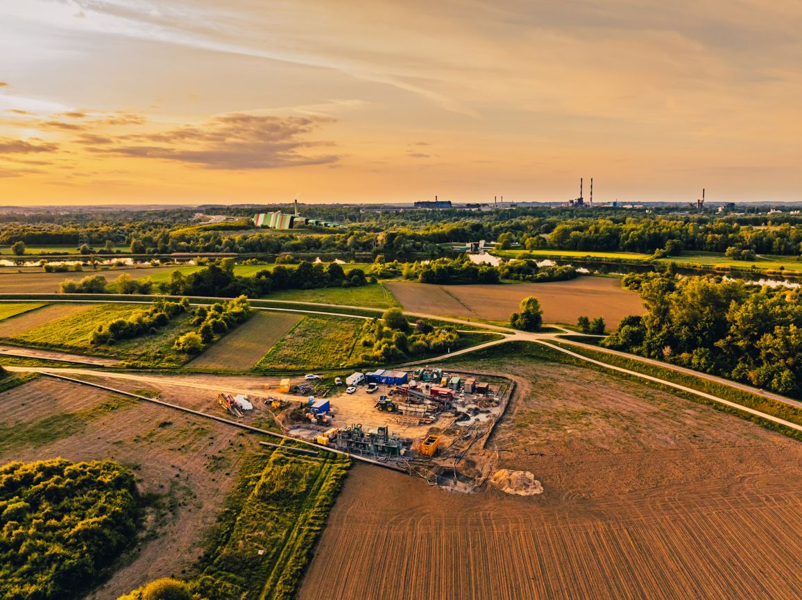 Zdjęcie lotnicze wykonane podczas złotego zachodu słońca, przedstawiające ogrodzony plac budowy z kontenerami i maszynami, zlokalizowany pośród pól uprawnych. W tle widać zakole rzeki, zielone lasy oraz industrialny horyzont z kominami fabrycznymi na tle ciepłego, pomarańczowego nieba.