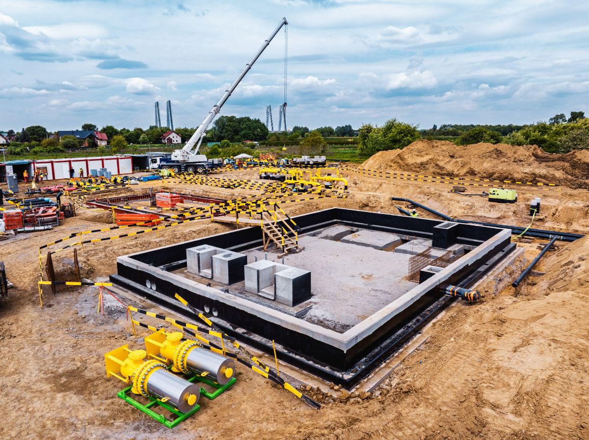 Widok z góry na głęboki wykop z dużym, prostokątnym betonowym fundamentem zabezpieczonym czarną izolacją. Na pierwszym planie, na zielonych konstrukcjach wsporczych, leżą dwa duże, masywne elementy armatury gazowej w kolorze srebrnym z żółtymi kołnierzami. W tle pracuje biały żuraw.