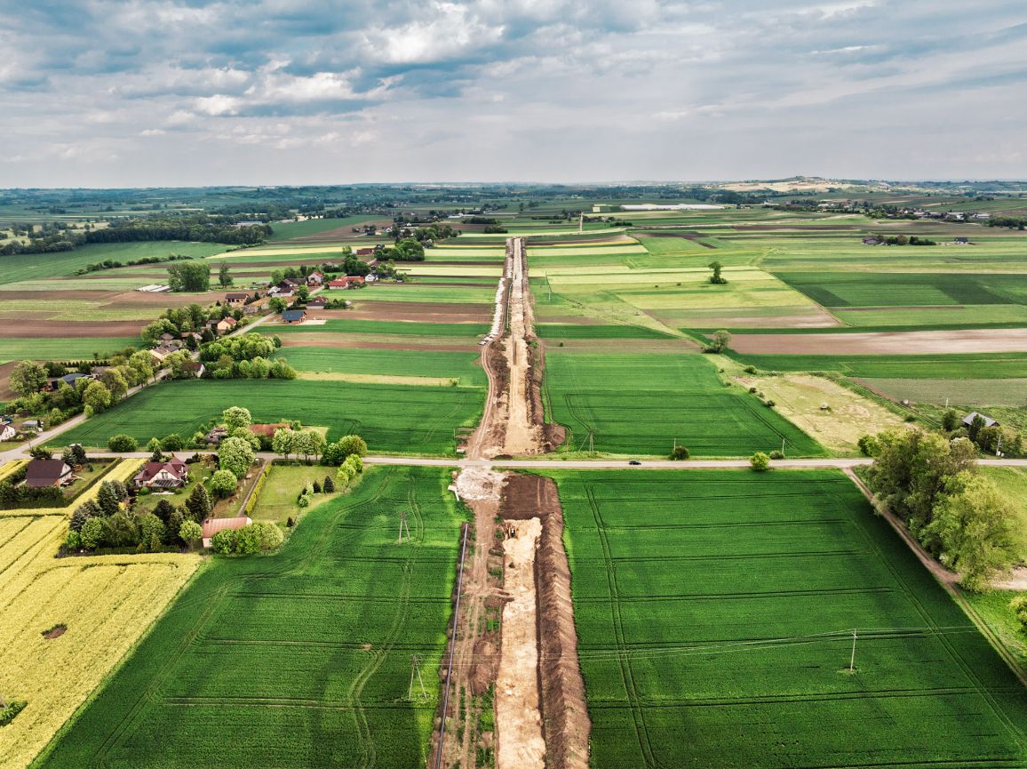 Szeroka panorama z lotu ptaka ukazująca rolniczy krajobraz z polami uprawnymi. Przez sam środek kadru, od dolnej do górnej krawędzi, biegnie długi, piaszczysty pas budowy gazociągu, który przecina prostopadle lokalną drogę asfaltową. Po lewej stronie widoczna jest niewielka wieś z domami jednorodzinnymi otoczonymi ogrodami.