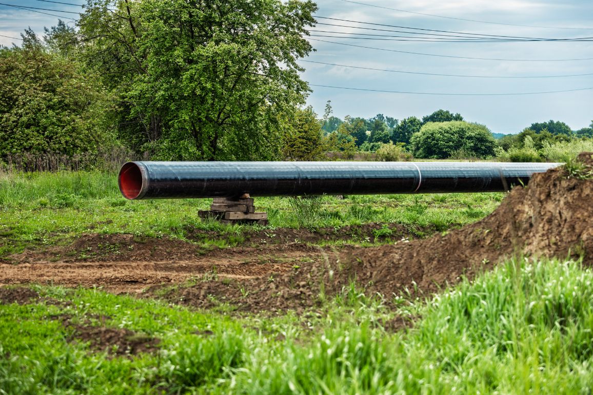 Gruba, stalowa rura gazociągowa z czarną powłoką izolacyjną i jaskrawoczerwonym wnętrzem ułożona poziomo na drewnianych podestach. Rura znajduje się na zielonej, trawiastej polanie, obok dużego nasypu ziemnego. W tle rosną gęste drzewa liściaste, nad którymi przebiegają przewody linii elektroenergetycznej.