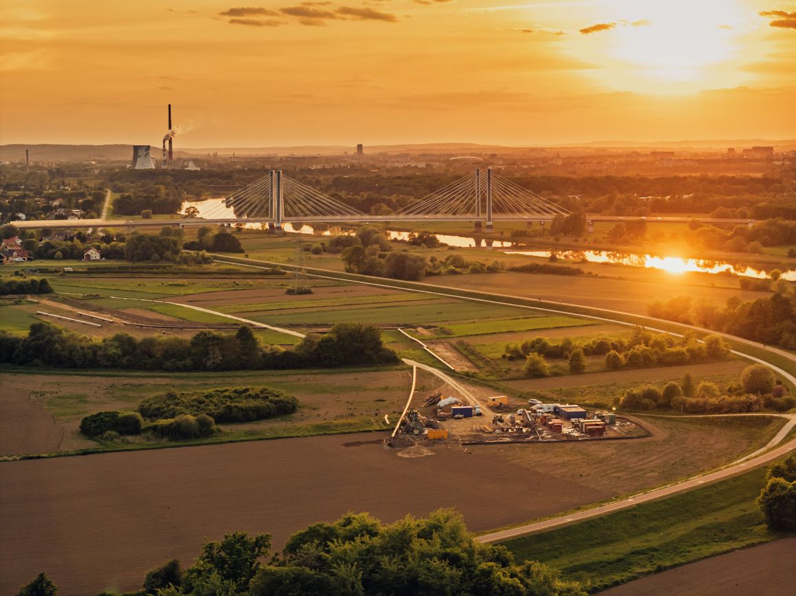 Panorama z lotu ptaka uchwycona pod słońce podczas malowniczego zachodu słońca. Na pierwszym planie widać bazę budowlaną na skraju zaoranego pola, a w tle szeroką rzekę, nad którą wznosi się duży, nowoczesny most wantowy (podwieszany). Na horyzoncie majaczy sylwetka elektrociepłowni.