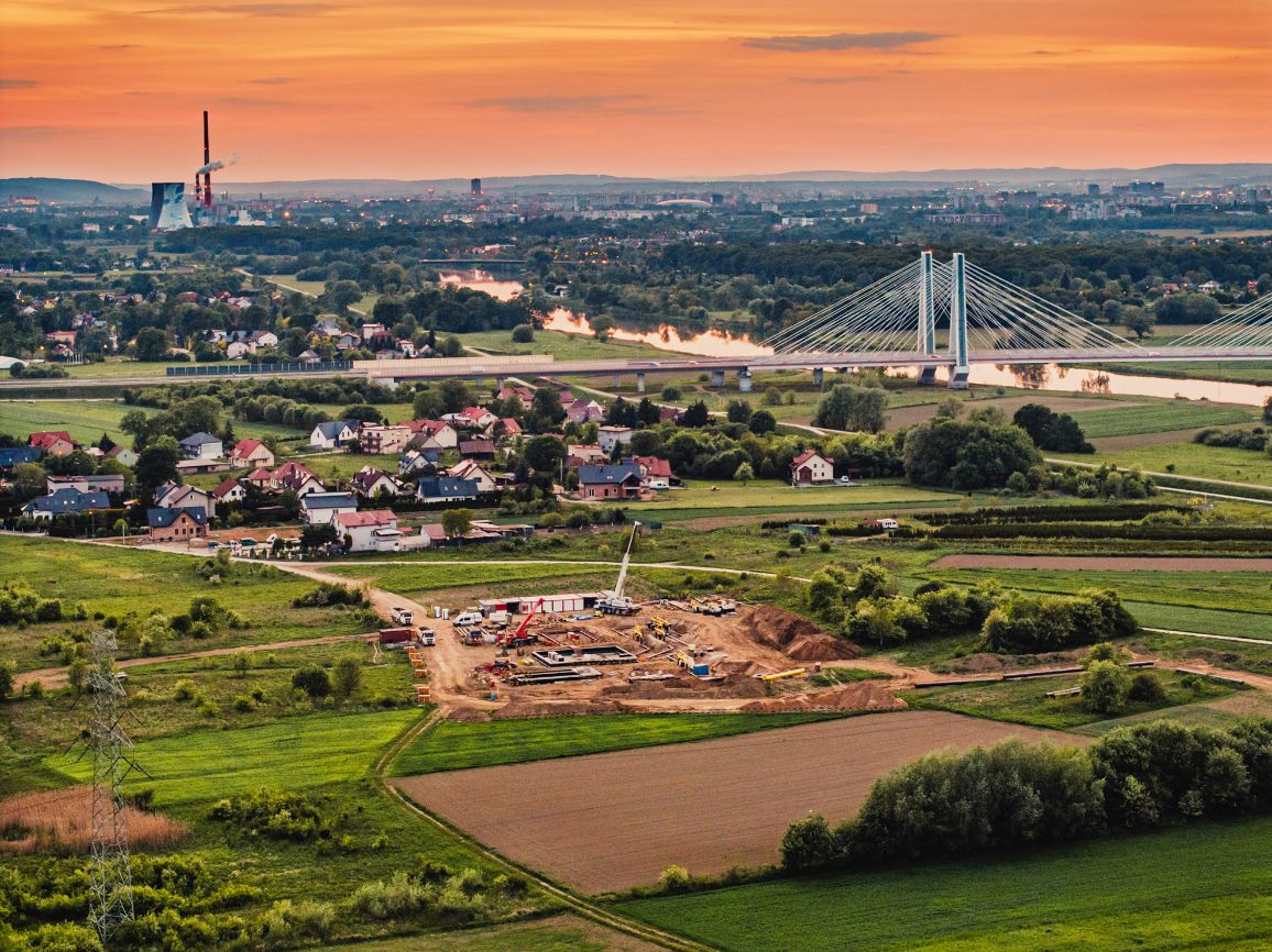 Zdjęcie lotnicze zrobione o zmierzchu, przedstawiające podmiejskie osiedle domów jednorodzinnych graniczące z polami uprawnymi. W dolnej części widoczny jest rozkopany plac budowy z dźwigiem, a w tle rzeka, długi podwieszany most drogowy oraz industrialna panorama miasta pod pomarańczowo-różowym niebem.