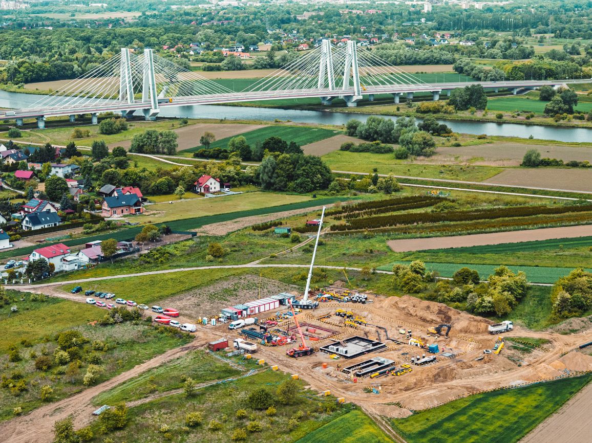 Zdjęcie z lotu ptaka przedstawiające duży plac budowy infrastruktury przesyłowej w ciągu dnia. W centralnej części widać betonowe fundamenty, głębokie wykopy, wysoki biały żuraw samojezdny, koparki oraz zaparkowane pojazdy. W tle widoczne są domy, rzeka i biały most podwieszany.