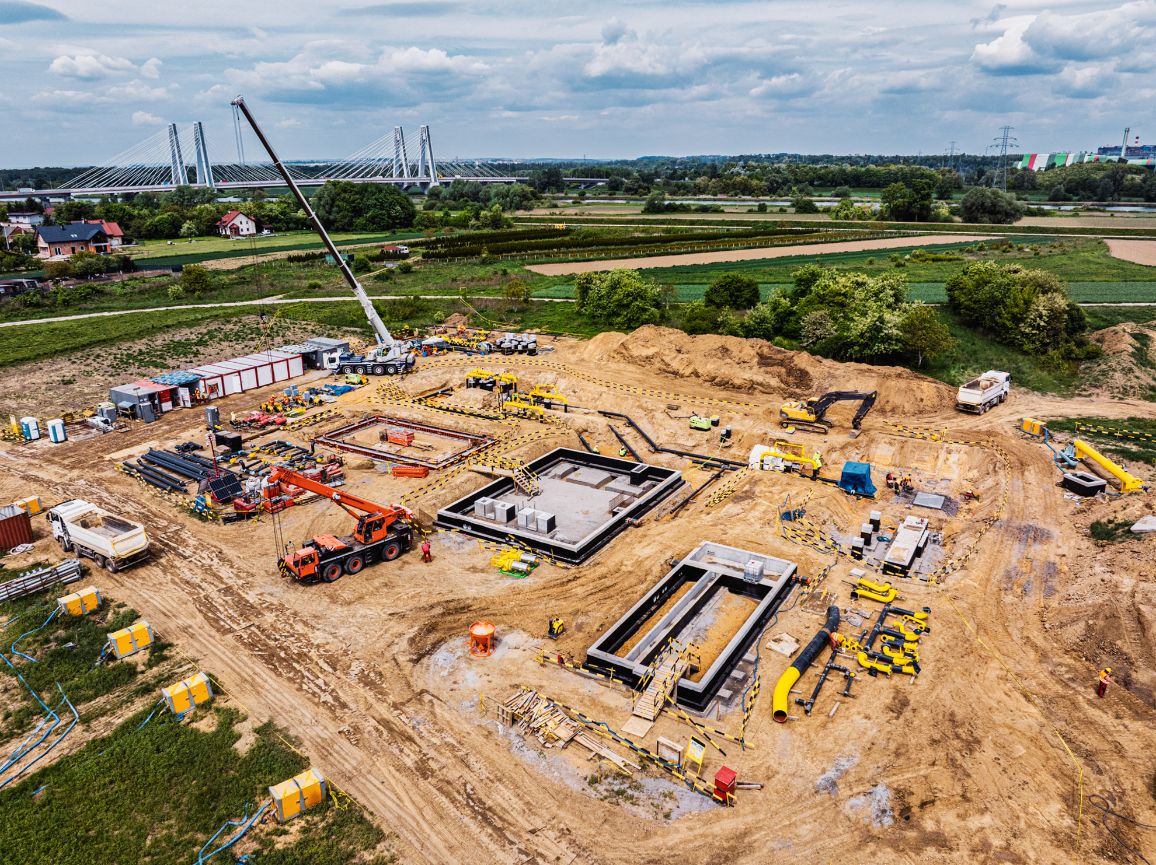 Średni kadr z lotu ptaka na zaawansowane prace ziemne i konstrukcyjne. Na piaszczystym terenie rozmieszczone są betonowe zbiorniki i fundamenty, pomarańczowy dźwig kołowy, żółta koparka oraz składowane materiały budowlane. Teren jest wygrodzony żółto-czarną taśmą ostrzegawczą.